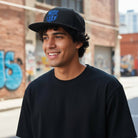 Young man wearing a black FC Barcelona Locale Snapback Hat with blue logo and flat brim outdoors in urban setting